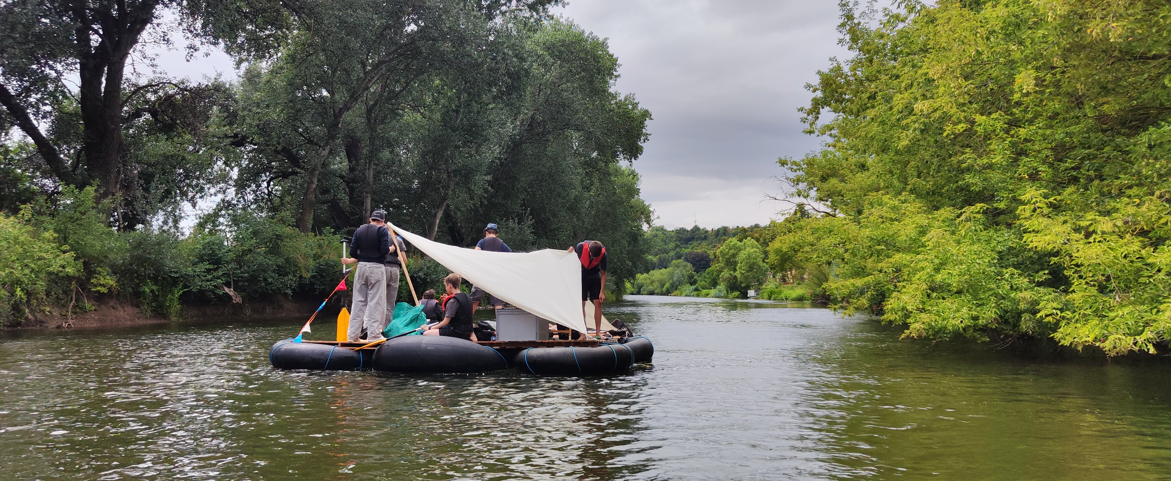 Ein Floß mit Menschen auf dem Fluss, Bäume an den Ufern.