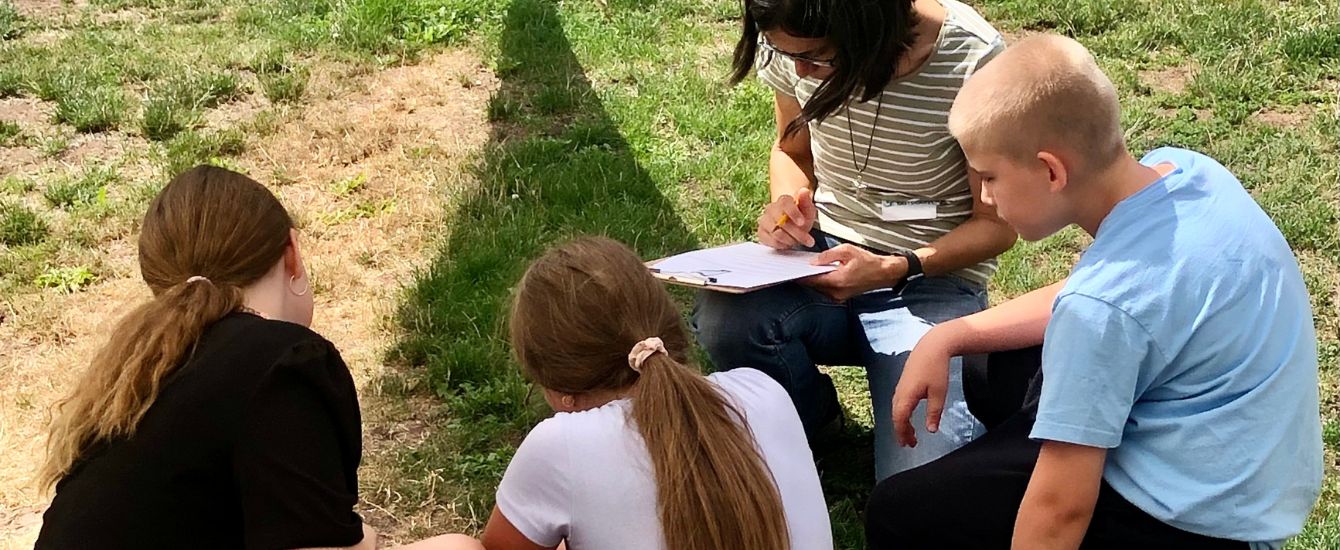 3 Kinder sitzen auf einer Wiese, eine Erwachsene Person schreibt auf einem Klemmbrett