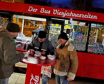 Drei Männer stehen vor dem umgebauten roten Reisebus des Vereins "Vierjahreszeiten" in Halle und nehmen an einem Stehtisch eine Mahlzeit zu sich.