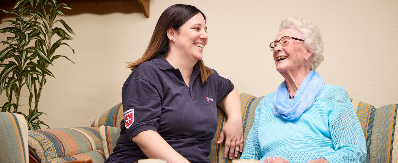 Maltesermitarbeiterin sitzt neben einer lachenden Rentnerin