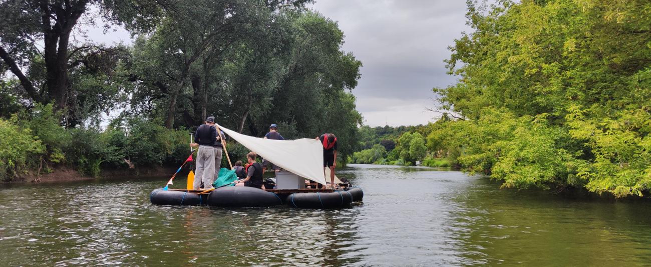 Ein Floß mit Menschen auf dem Fluss, Bäume an den Ufern.