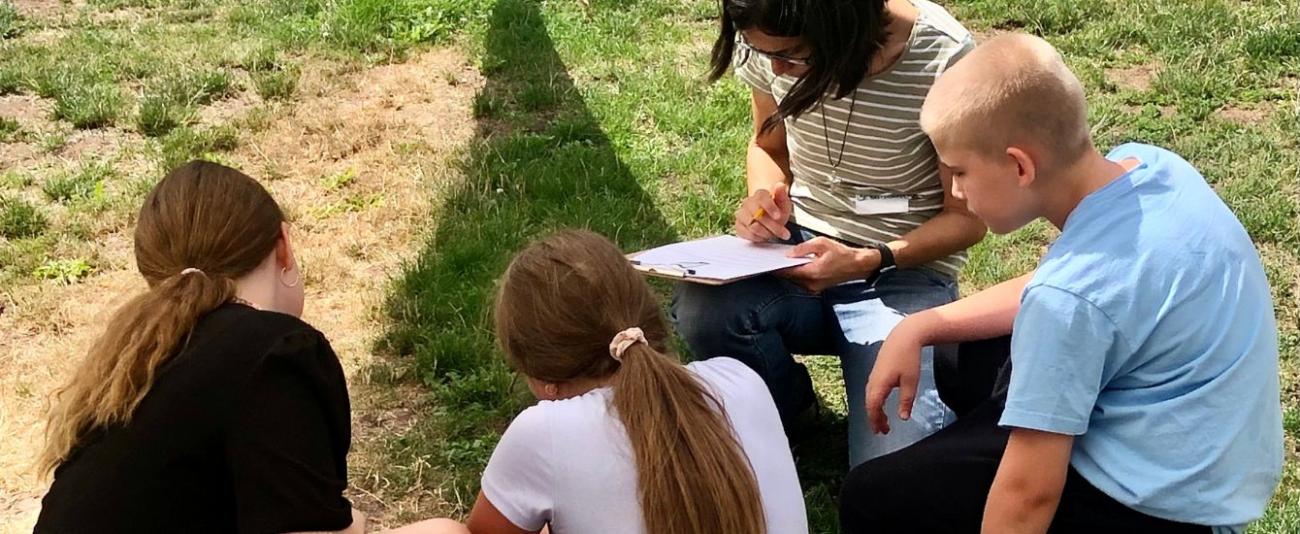 3 Kinder sitzen auf einer Wiese, eine Erwachsene Person schreibt auf einem Klemmbrett