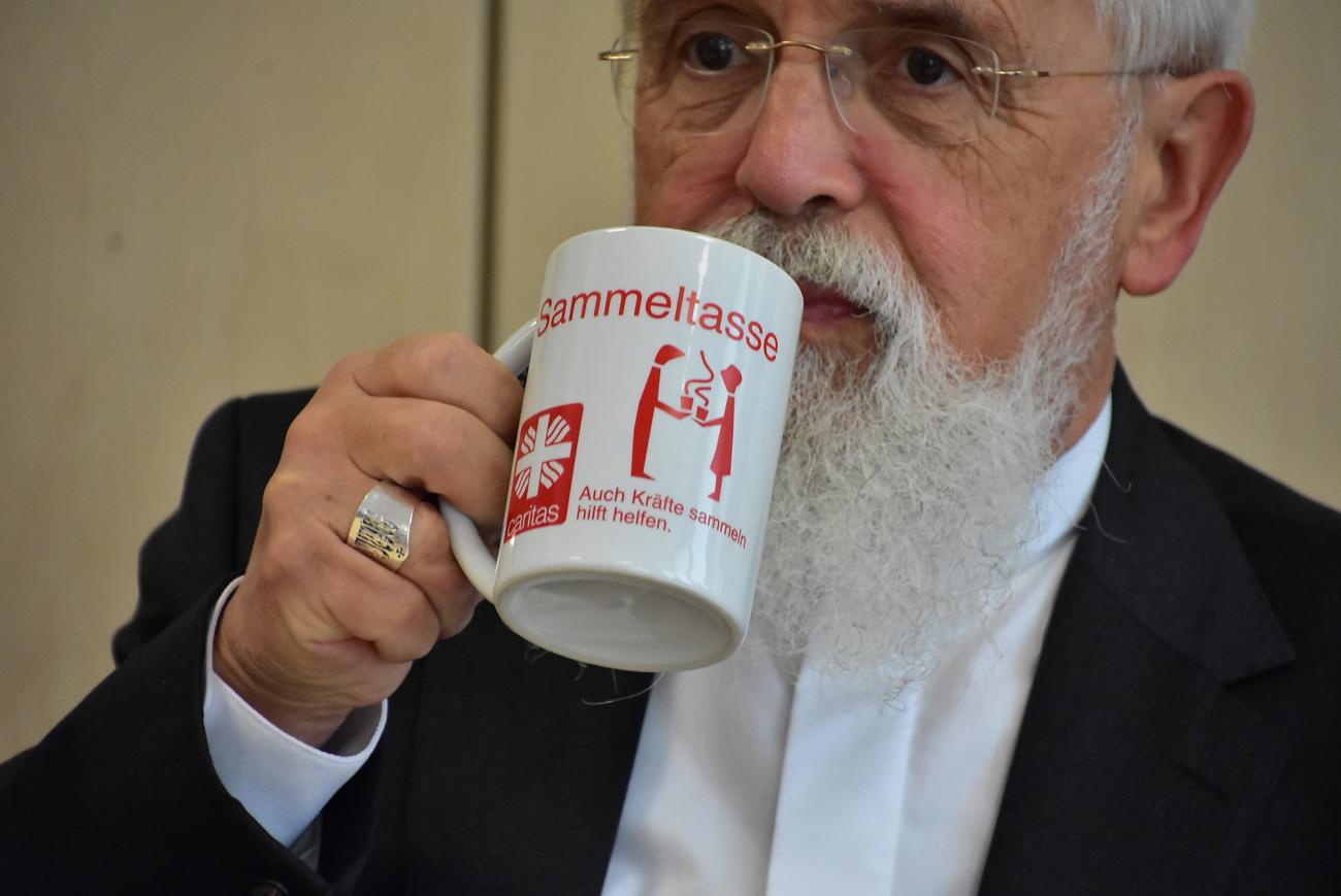 Bischof Feige trinkt aus einer Caritas-Tasse mit der Aufschrift "Sammeltasse"