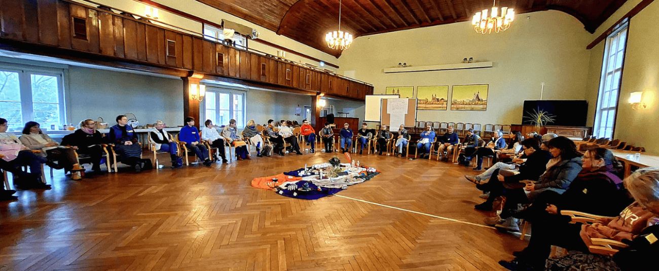 Eine etwa 50-köpfige Gruppe von Menschen sitzt im Stuhlkreis in einem großen Saal